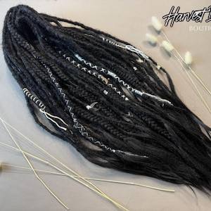 May include: Black dreadlocks with silver and white accents, styled with decorative wraps and beads. The image includes the text "Harvest Dreads Boutique" in a script font, and is decorated with dried flowers.