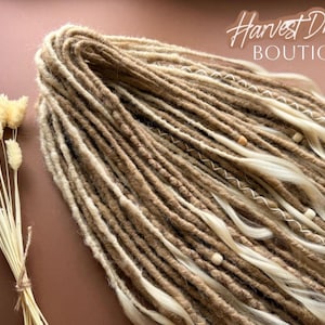 a collection of long, braided hair strands in various shades of brown and blonde, arranged on a pink background. There are also some dried flowers visible in the image.