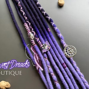 May include: Close-up of purple and blue dreadlocks with decorative silver charms. The dreadlocks are adorned with skull, bat, and sun-shaped charms. The text "Harvest Dreads Boutique" is visible in the image.