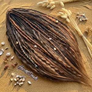 May include: A set of brown and blonde synthetic dreadlocks featuring various textures, including braids, twists, and loose wavy strands. The hair extensions are adorned with small white shell beads and white thread wraps. The text Harvest Dreads is visible.