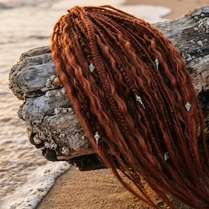Könnte beinhalten: Ein Set aus kastanienbraunen synthetischen Dreadlocks mit einer Mischung aus welligen und geflochtenen Texturen. Die Haarverlängerungen sind mit kleinen, silberfarbenen Metallanhängern in Form von Totenköpfen und geometrischen Formen verziert.