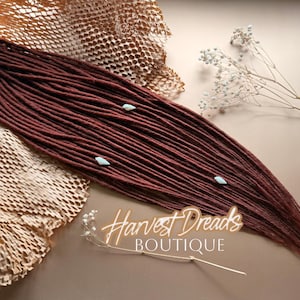 May include: A bundle of burgundy dreadlocks with small, light blue accents. The dreadlocks are arranged on a neutral surface with decorative paper and sprigs of white flowers. The text "Harvest Dreads Boutique" is visible.