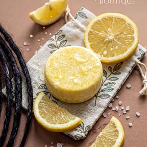 May include: A round yellow shampoo bar sits on a beige drawstring pouch with a leaf pattern, surrounded by lemon slices and scattered salt crystals. Black dreadlocks are visible on the left. The text Harvest Dreads BOUTIQUE is at the top.
