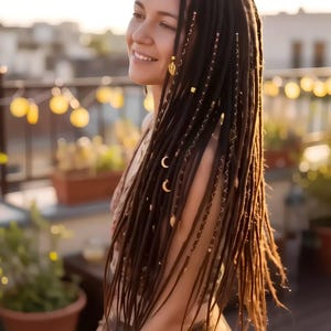 Könnte beinhalten: Lange dunkelbraune Dreadlocks, verziert mit goldfarbenen Metall-Haaraccessoires, darunter kleine Mondsichel-Anhänger, blattförmige Anhänger und dünne, um die Strähnen gewickelte Metallfäden. Die Haarverlängerungen sind locker gestylt.