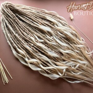 "Handmade in USA sand and vanilla butter blonde synthetic crochet dreadlocks with loose ends and wavy curls by Harvest Dreads Boutique. The set features textured boho dreads decorated with milk chocolate thread wraps and natural wooden beads on a warm background." 🌾