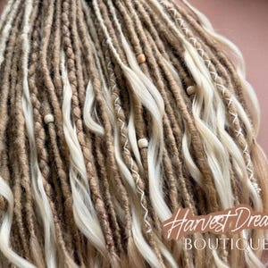 a close-up view of a collection of braided hair strands in various shades of brown and white, with small white beads scattered throughout. The image also includes a small white flower and the text "Harvest Dresses Boutique" in the background.