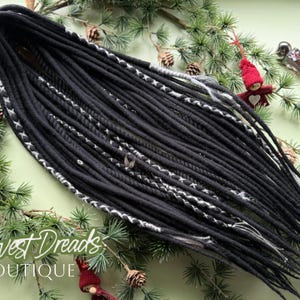 Puede incluir: Dreadlocks negros con detalles blancos y grises, dispuestos sobre un lecho de agujas de pino y piñas. El texto "Harvest Dreads Boutique" es visible. Los dreadlocks son de un color oscuro y sólido.