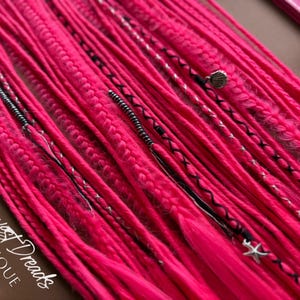May include: Bright pink dreadlocks with decorative accents. The dreads are adorned with silver beads, black thread, and seashell and starfish charms. The Harvest Dreads Boutique logo is visible in the lower left corner.