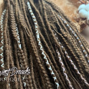 May include: Close-up of brown dreadlocks with white and pink accents. The dreads are neatly styled and appear to be handmade. The text "Harvest Dreads BOUTIQUE" is visible in the lower left corner. The background is a neutral colour.
