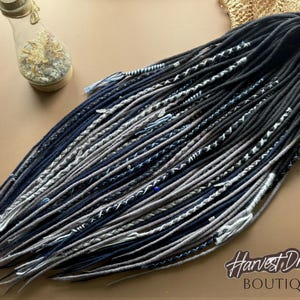 May include: A set of long, thin hair extensions in shades of black, dark blue, and light gray. The strands feature various textures, including smooth, braided, and wrapped sections with small decorative beads and thread accents throughout.