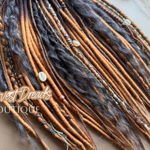 May include: Close-up of a collection of dreadlocks in shades of brown, orange, and gray. Some dreads are adorned with small shells and beads. The text "Harvest Dreads BOUTIQUE" is visible.