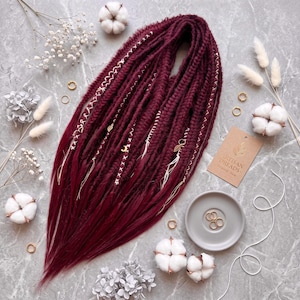 May include: A set of deep burgundy synthetic dreadlocks featuring braided textures, wrapped accents, and small gold-toned charms including a crescent moon and leaf. A tan tag labeled Artisan Dreads is present alongside a small gray dish with gold rings.