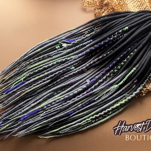 May include: A set of synthetic dreadlocks in shades of black, charcoal gray, and lime green. The strands feature decorative accents including braided sections, thin colored thread wraps, and small metallic beads throughout the hair extensions.