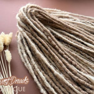 a close-up view of a bundle of brown and white braided hair, with some dried flowers next to it. The background is a pinkish-brown color.
