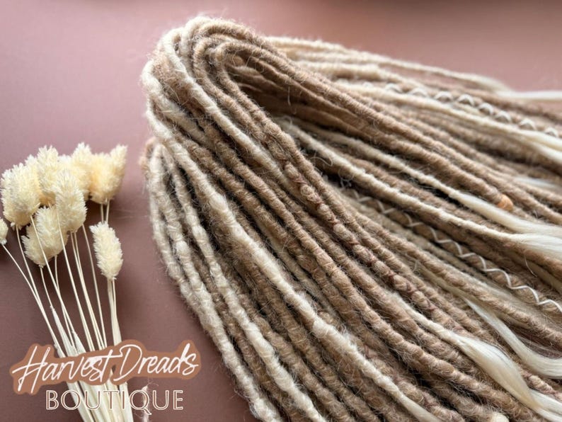 a close-up view of a bundle of brown and white braided hair, with some dried flowers next to it. The background is a pinkish-brown color.