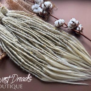 Puede incluir: Dreadlocks rubios sobre una superficie marrón, con flores de algodón. Los dreadlocks son de un rubio claro natural, con raíces ligeramente más oscuras. El texto "Harvest Dreads Boutique" es visible.