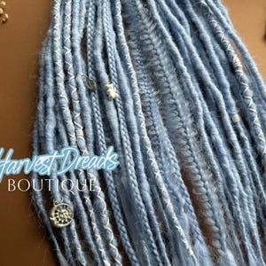 Könnte beinhalten: Nahaufnahme von blauen Dreadlocks mit weißen Nähten und dekorativen Charms. Die Dreadlocks sind in einem Bündel angeordnet, mit dem Text "Harvest Dreads Boutique" in hellblauer Schrift.