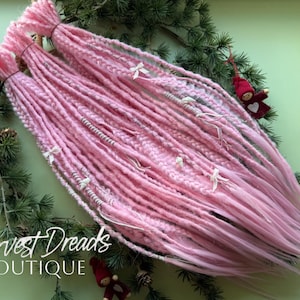 Puede incluir: Dreadlocks rosas con lazos blancos y detalles plateados sobre ramas de pino. El texto "Harvest Dreads Boutique" es visible. Pequeños adornos rojos están dispersos alrededor de los dreadlocks.
