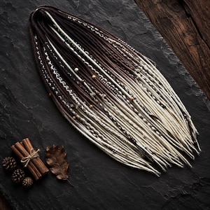 May include: A set of synthetic dreadlocks featuring a color gradient that transitions from dark brown at the roots to a light cream at the tips. The dreadlocks are adorned with various decorative wraps, small wooden beads, and metallic accents.