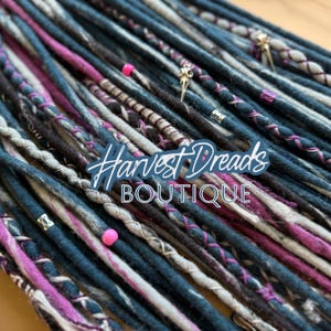 Könnte beinhalten: Nahaufnahme von Dreadlocks in den Farbtönen Teal, Grau, Schwarz und Pink. Einige Dreadlocks sind mit dekorativen Perlen und Charms verziert. Der Text "Harvest Dreads BOUTIQUE" ist sichtbar.