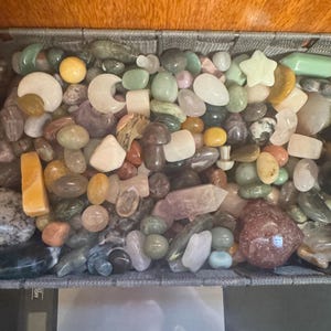 Puede incluir: Una colección de piedras preciosas y cristales pulidos de diversas formas, incluyendo lunas, estrellas, setas y puntas, contenidos en una cesta tejida gris. Las piedras presentan una paleta de colores variada como verde, rosa, amarillo y transparente.