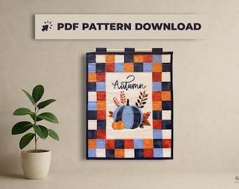 May include: A quilted wall hanging with a fall theme. The design features a blue pumpkin with orange accents, the word "Autumn", and a border of blue, orange, and white squares. The text "PDF PATTERN DOWNLOAD" is above the wall hanging.