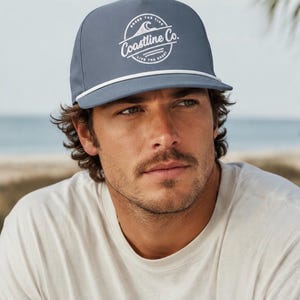 Op de afbeelding: Een leigrijze baseballpet met een wit koorddetail langs de rand en een rond wit grafisch logo op de voorkant. Het logo toont een golfpictogram en de tekst Coastline Co. met de zinnen Chase The Tide en Live The Coast.
