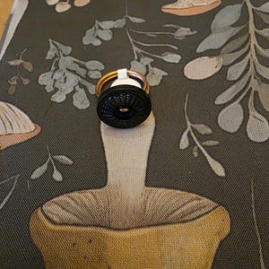 May include: A black bottle stopper with a textured design, featuring a gold-colored ring and a white band. The stopper is set against a dark green fabric with a mushroom and leaf pattern.