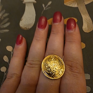 May include: A gold-colored ring with a detailed embossed design. The ring is circular and appears to be made of metal. The ring is worn on a finger with red nail polish. The background features a mushroom pattern.
