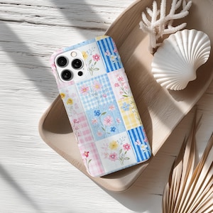 Pode incluir: Capa de telefone com padrão floral de patchwork em cores pastel. O design inclui quadrados de xadrez rosa, xadrez azul e listras amarelas, adornados com delicadas ilustrações de flores. A capa está em uma bandeja de madeira com uma concha e coral.