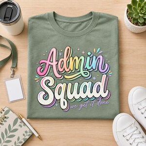 May include: An olive green t-shirt featuring a colorful graphic with the text "Admin Squad" in a bold, layered, multi-colored font, followed by the smaller script text "we get it done" and several small star and sparkle accents.