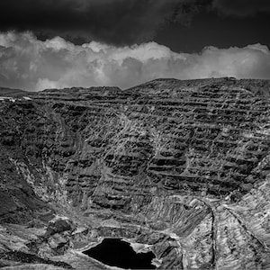May include: Black and white landscape photograph of a deep crater with layered rock formations. A dark lake sits at the bottom, and the sky is filled with dramatic, dark clouds. The image captures a sense of depth and scale.
