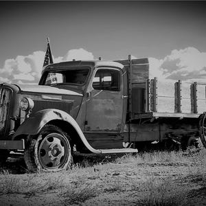 May include: Black and white image of an antique flatbed truck with an American flag. The truck is weathered and parked in a field. The sky is cloudy.