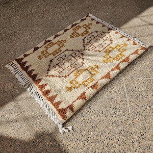 May include: A rectangular, cream-colored rug with a geometric pattern in shades of brown and gold. The rug has a fringed edge and is placed on a stone surface. The design features repeating shapes and lines.