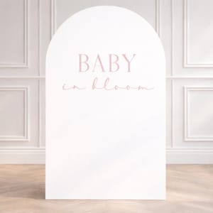 May include: A white, arched sign with the text "BABY in bloom" in a soft pink hue. The sign is placed in front of a white wall with decorative molding and a wooden floor.