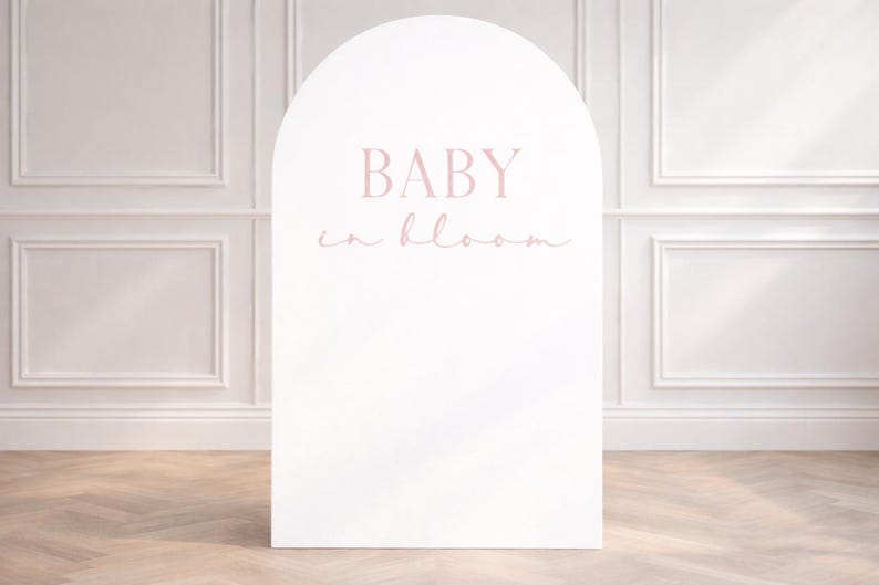 May include: A white, arched sign with the text "BABY in bloom" in a soft pink hue. The sign is placed in front of a white wall with decorative molding and a wooden floor.