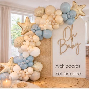 May include: A baby shower decoration featuring a white arch with a woven texture, adorned with balloons in shades of blue, white, and beige. Gold star and seashell-shaped balloons add a nautical touch. The words "Oh Baby" are displayed in gold script.