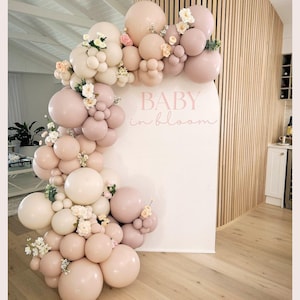May include: A decorative balloon arch in pink and cream hues, decorated with small white flowers, curves around a white backdrop. The words "BABY in bloom" are written in pink script. The arrangement is set against a wooden slatted wall.