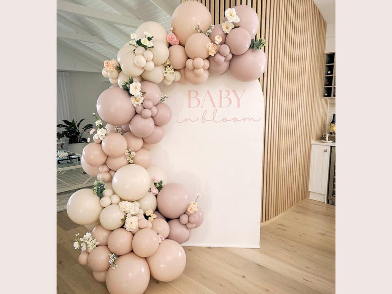 May include: A decorative balloon arch in pink and cream hues, decorated with small white flowers, curves around a white backdrop. The words "BABY in bloom" are written in pink script. The arrangement is set against a wooden slatted wall.