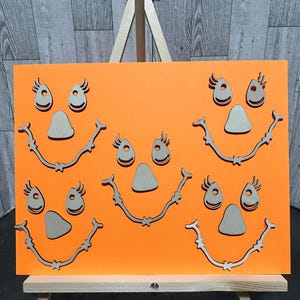 Op de afbeelding: Een oranje Halloween-bord met meerdere lachende pompoengezichten. Elk gezicht heeft uitgesneden ogen, neuzen en monden van hout. Het bord wordt gepresenteerd op een kleine houten ezel, ideaal voor seizoensdecoratie.
