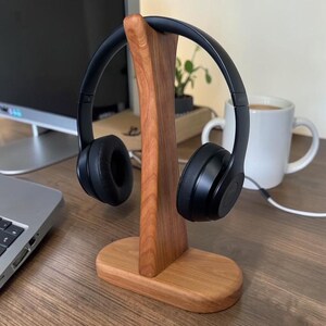 May include: A pair of black over-ear headphones resting on a smooth, light brown wooden stand. The stand features a vertical support post attached to an oval-shaped base, designed to hold the headphones securely in an upright position.