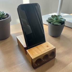 Puede incluir: Un soporte de teléfono de madera con un acabado natural. El soporte tiene una base rectangular con tres agujeros circulares y una ranura para sostener un teléfono inteligente. Un teléfono inteligente negro se coloca en el soporte, inclinado ligeramente hacia arriba.