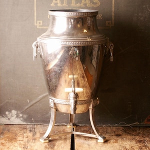 May include: A vintage silver-plated coffee urn with a spigot and ornate detailing. The urn has a large, bulbous body and stands on three curved legs. The background features text that reads "OFFICIAL QUARTER SECTIONAL ATLAS".
