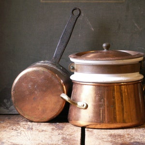 Może przedstawiać: Dwa miedziane garnki z ciemnymi metalowymi uchwytami i miedziany i biały garnek do kąpieli wodnej. Garnki stoją na drewnianej powierzchni. Miedź ma czerwonobrązowy kolor, a uchwyty są ciemnoszare.