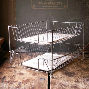 May include: A vintage two-tier wire desk organizer. The organizer is made of silver-colored metal and holds several old papers. The background features text that reads "Official Quarter Sectional Atlas of the City of Milwaukee."
