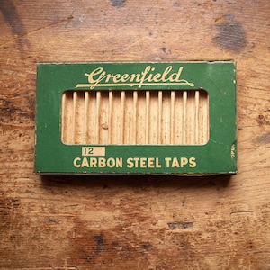 May include: A vintage green box of Greenfield carbon steel taps, with the number 12 visible. The box has a rectangular opening revealing the taps. The text "Greenfield" is in a script font.