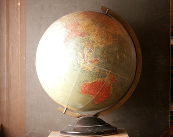 Puede incluir: Un globo terráqueo antiguo con un mapa detallado, montado sobre un soporte de metal oscuro, descansa sobre dos libros antiguos. El globo presenta tonos de verde, marrón y rojo, sobre un fondo oscuro.