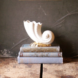 May include: White ceramic seashell-shaped vase with gold accents, sitting on a stack of three vintage books with blue covers.