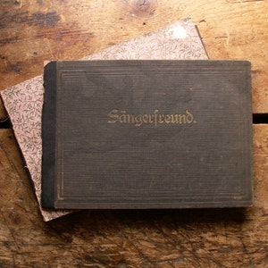 May include: A vintage book with the word "Sängerfreund" in gold lettering on the cover, resting on a patterned paper. The book is dark gray with a black spine. The background is a rustic wooden surface.
