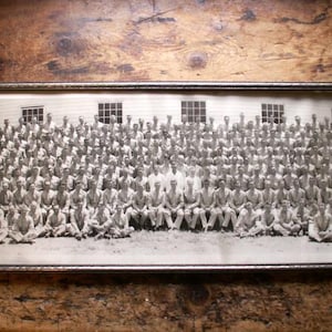 May include: A large, rectangular, black and white panoramic photograph framed in a thin metallic border. The image depicts a massive group of individuals posed in rows in front of a building with several windows, captured in a wide, horizontal format.